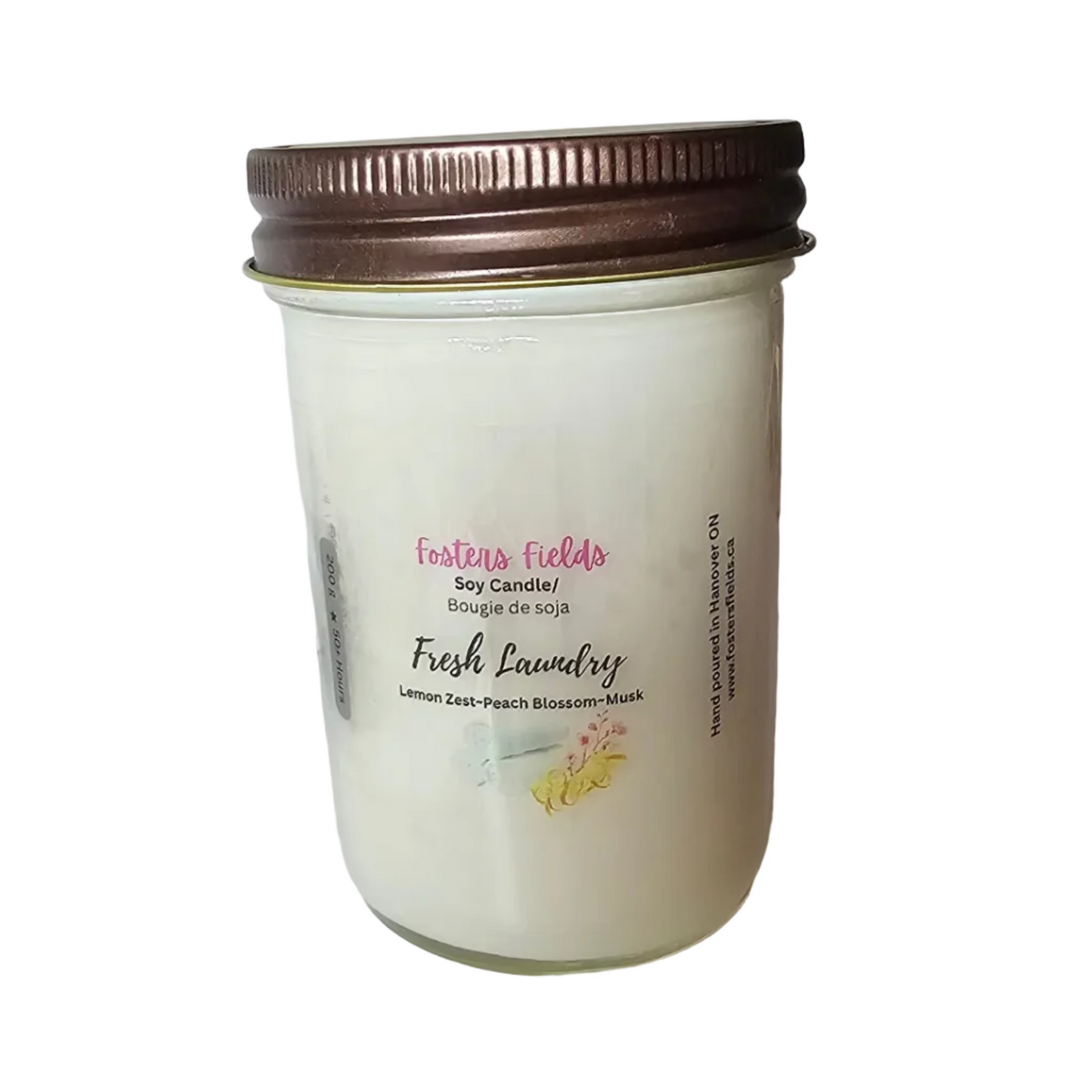 A jar candle labeled 'Fresh Laundry' by Fantasy Fabrics sits on a white shelf. The candle has an off-white wax body, a brown metal screw-top lid, and a white label with pink and black text.