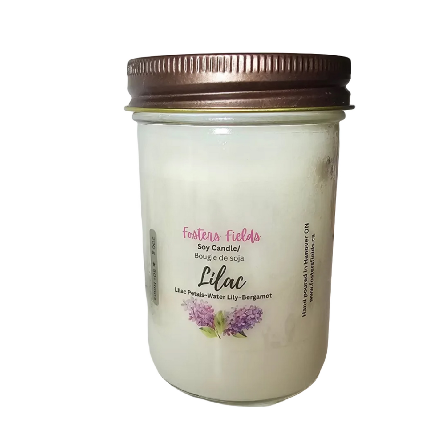 A jar candle with white wax and a metallic bronze lid sits on a white shelf. The front label reads 'Coconut Fields' and 'Lilac' with a small purple flower illustration.