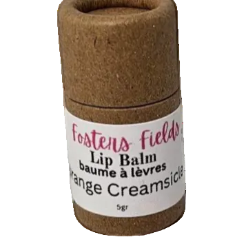 A cylindrical container of Orange Creamsicle Lip Balm with a label that reads 'Foster's Field Lip Balm Orange Creamsicle 5g'. The container appears to be made of cardboard or a similar lightweight material.