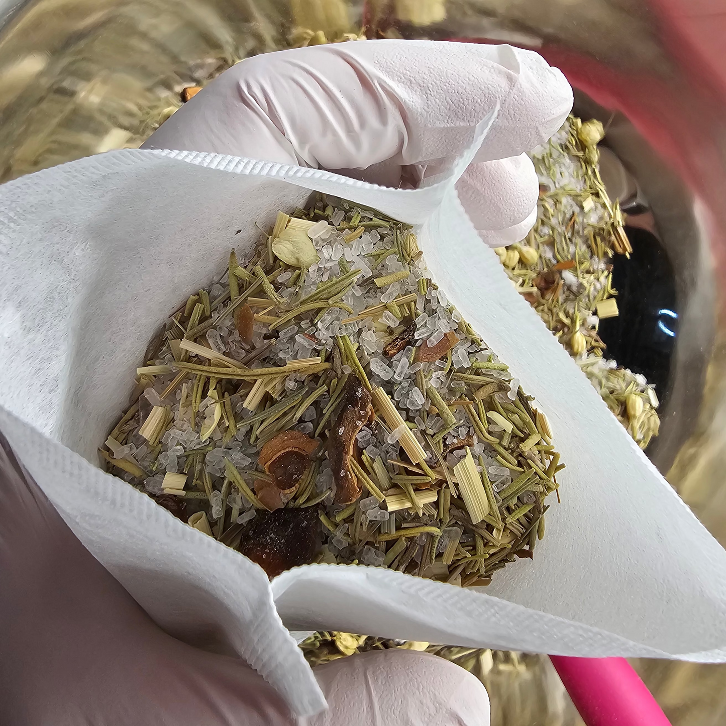 Hand wearing a white glove holding a triangular tea infuser filled with herbs working over a glass bowl. SHowing the creation of the product