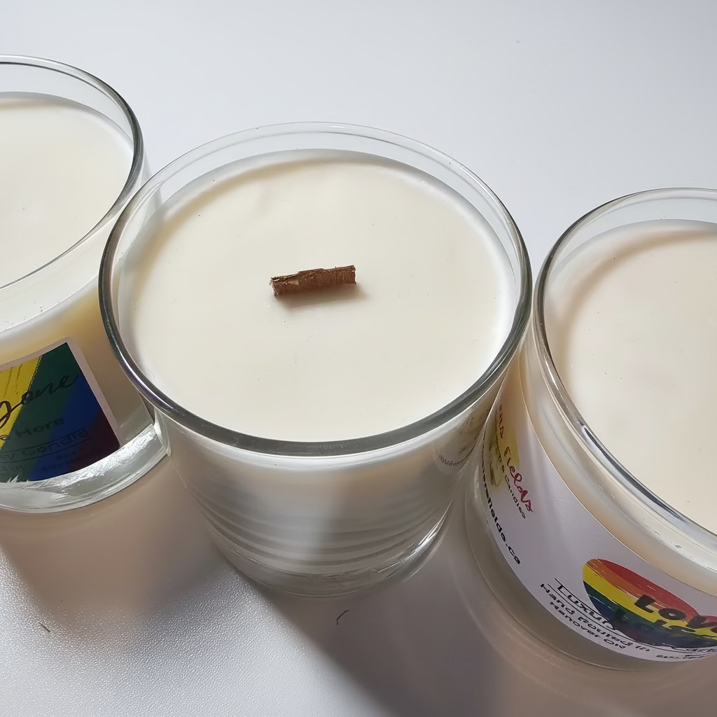 Three candles in glass jars on a white surface with visible labels. Rainbow pride labels