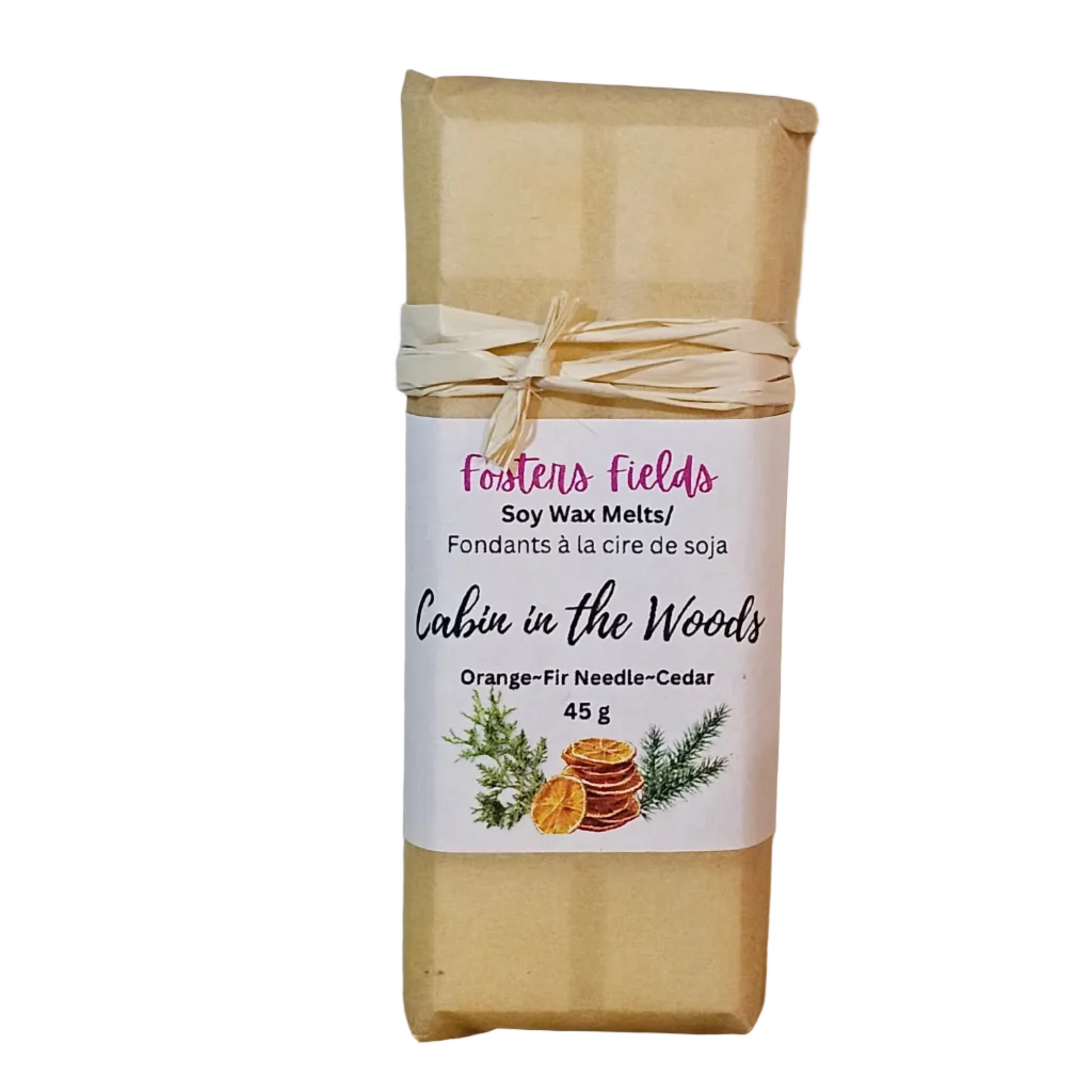 A wrapped bar of soy wax melt from Fosters Fields, labeled 'Cabin in the Woods' (Orange-Fir Needle-Cedar), wrapped in brown kraft paper with a beige twine bow and a white label, standing on a wooden surface against a plain wall.