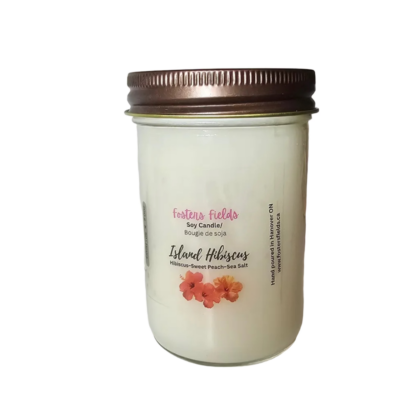 A jar candle labeled 'Fantasy Lights' and 'Island Hibiscus' with a metallic bronze lid, sitting on a white shelf next to decorative driftwood.